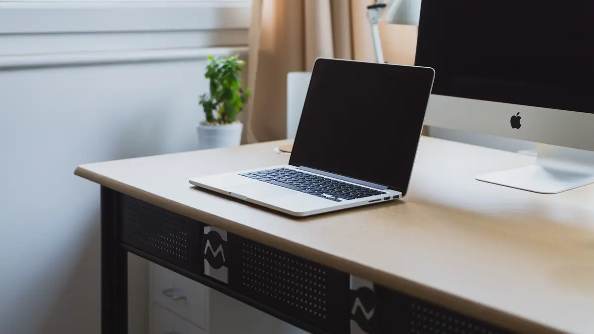 Notebook plateado abierto sobre un escritorio blanco junto a una ventana y una planta